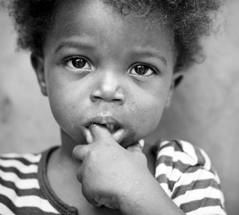 Black child with curly hair in a striped shirt, looking thoughtfully at the camera.